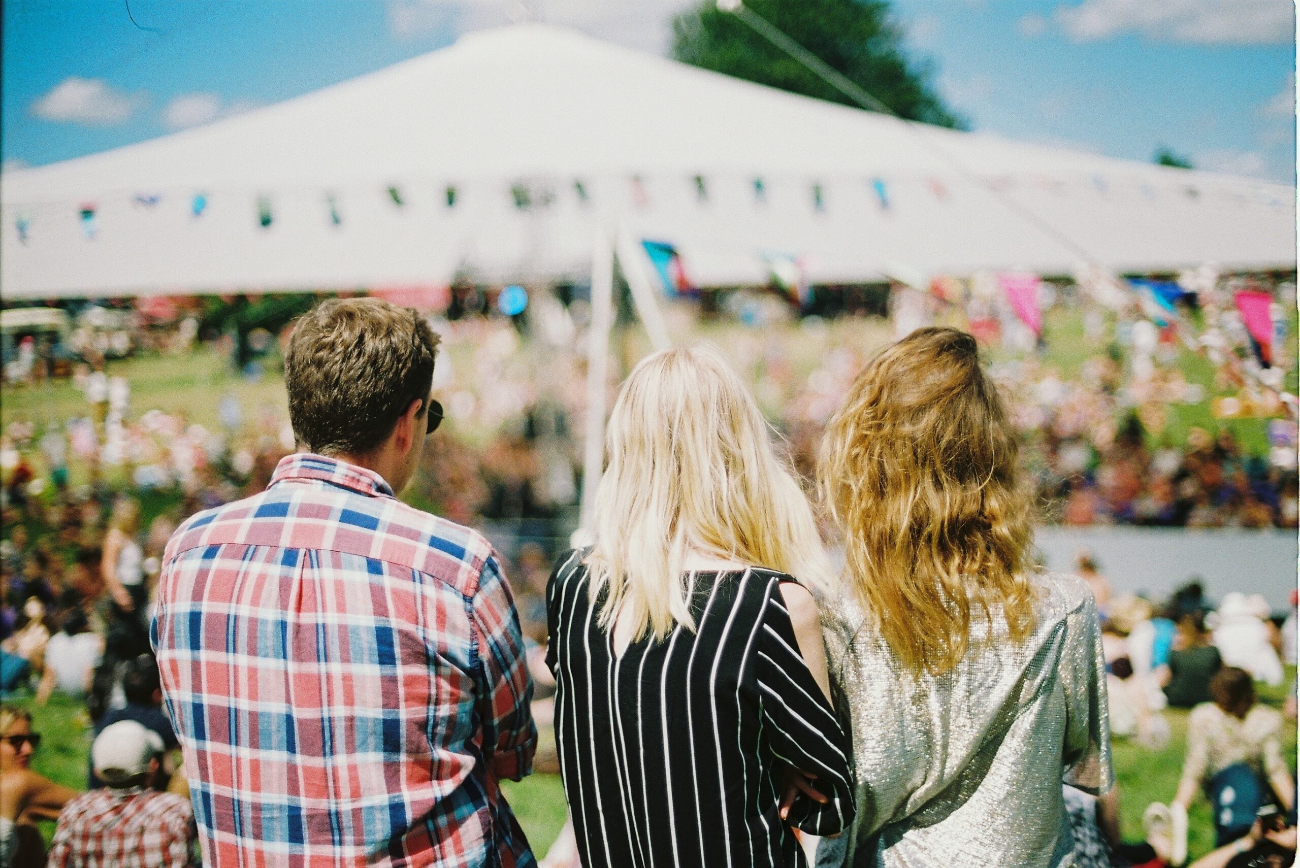 backs of three people sat in front of big tent at a festival.