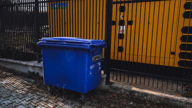 blue four wheel bin in front of a metal fence.